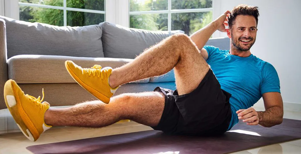 A smiling man in a blue shirt and yellow shoes performs an ab exercise on a mat in a bright living room.