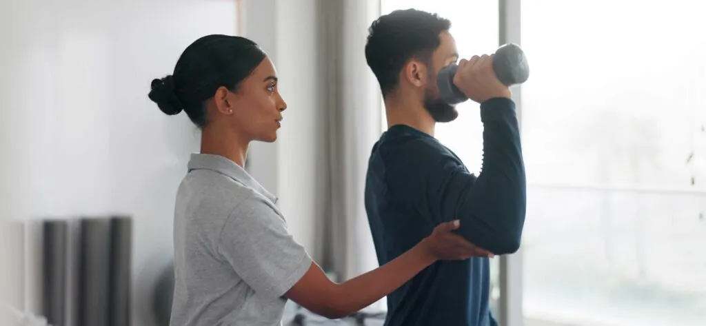 A female therapist assists a man performing a bicep curl with a dumbbell.