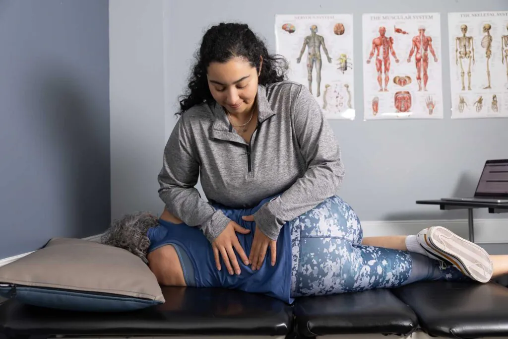 A practitioner in a grey top adjusts the lower back of a patient lying on their side on an examination table, with anatomical charts in the background.