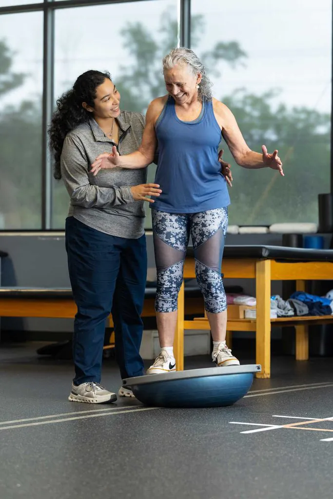 A physical therapist assists an older woman standing on a blue BOSU ball for balance training in a gym.
