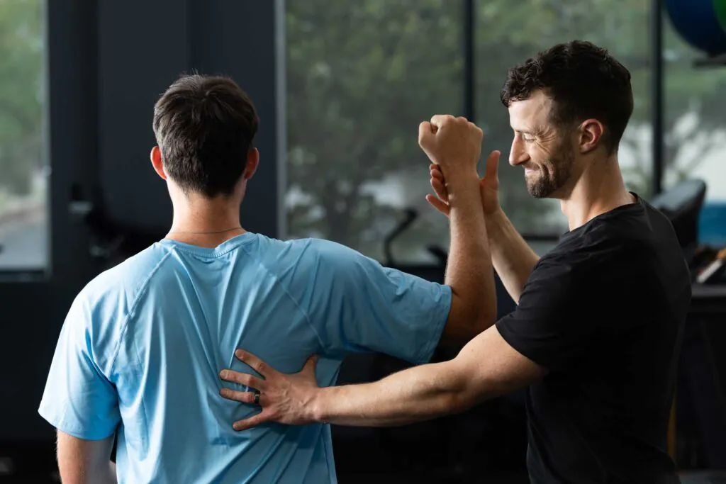 A man in a black shirt assists another man in a blue shirt with an arm and back exercise in a gym setting.