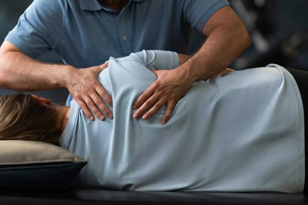 A practitioner's hands are on a patient's upper back, who is lying down on an examination table.