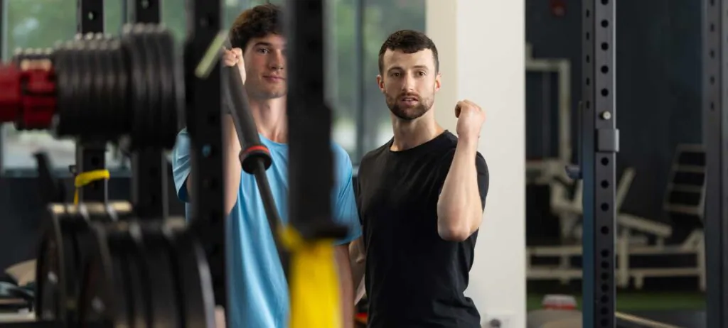 Two men in a gym, one holding a barbell and the other flexing his arm.