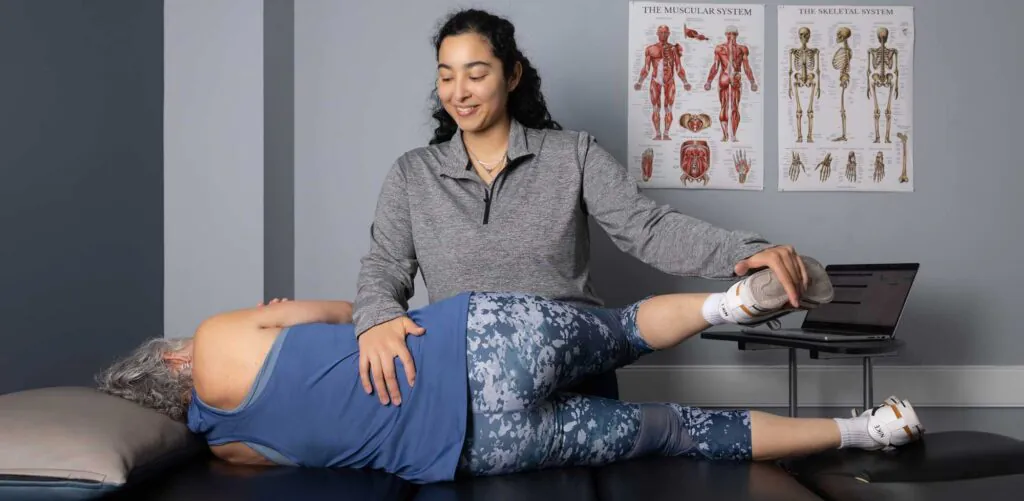 A physical therapist helps a patient with a leg stretch on an examination table, with anatomical charts in the background.