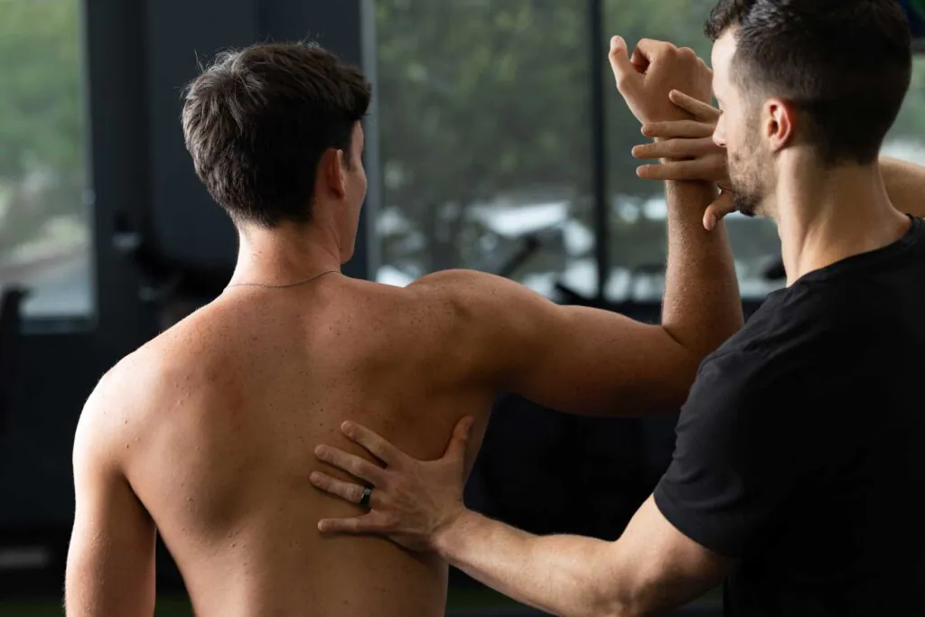Two men. One shirtless man from behind, receiving an arm and back adjustment from another man in a black t-shirt.