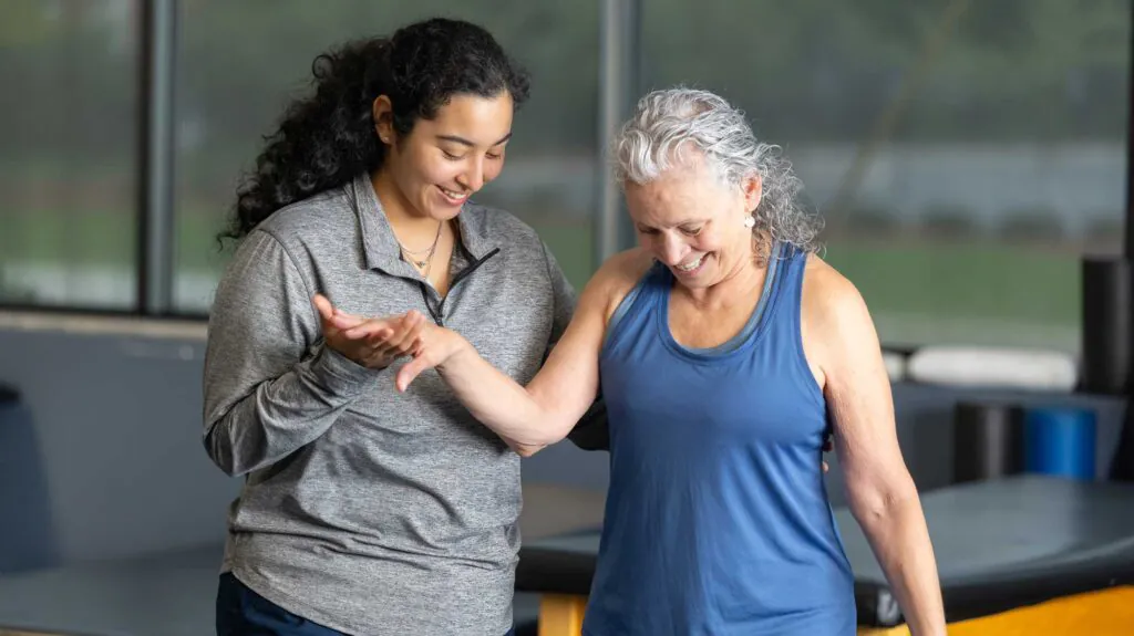 A young female physical therapist helps an older woman with an arm exercise, both smiling.