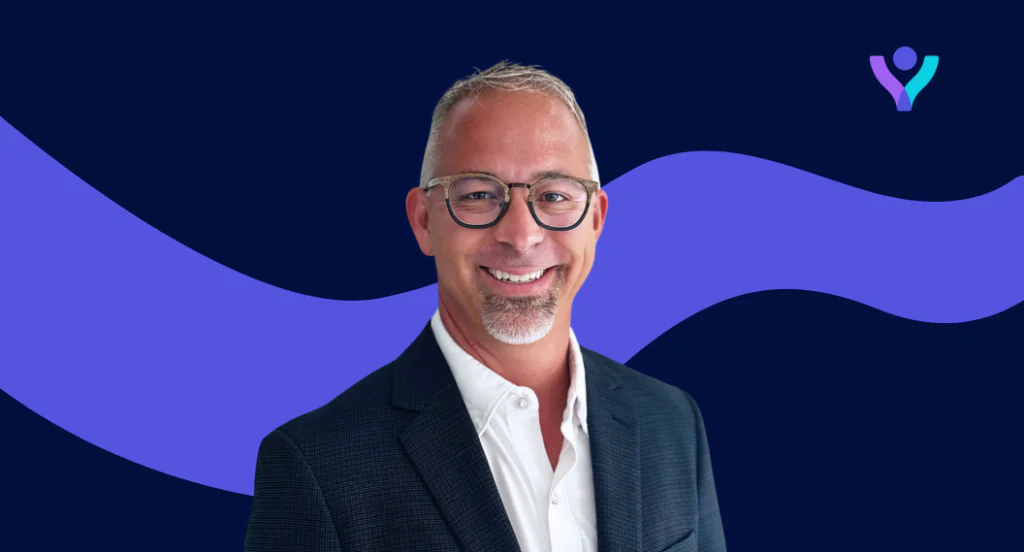 Headshot of a smiling man with gray hair, glasses, and a goatee, wearing a dark suit jacket over a white shirt. The background is dark blue with wavy purple accents and a small logo in the top right.
