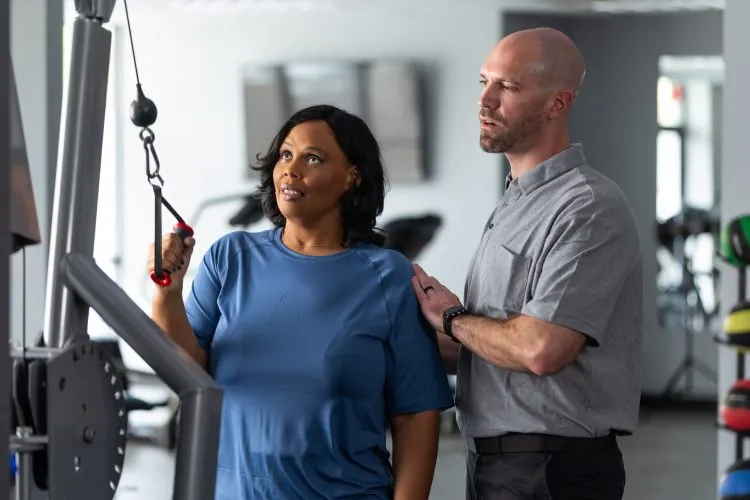 a black female patient being aided with a pull down weight exercise with a physical therapist