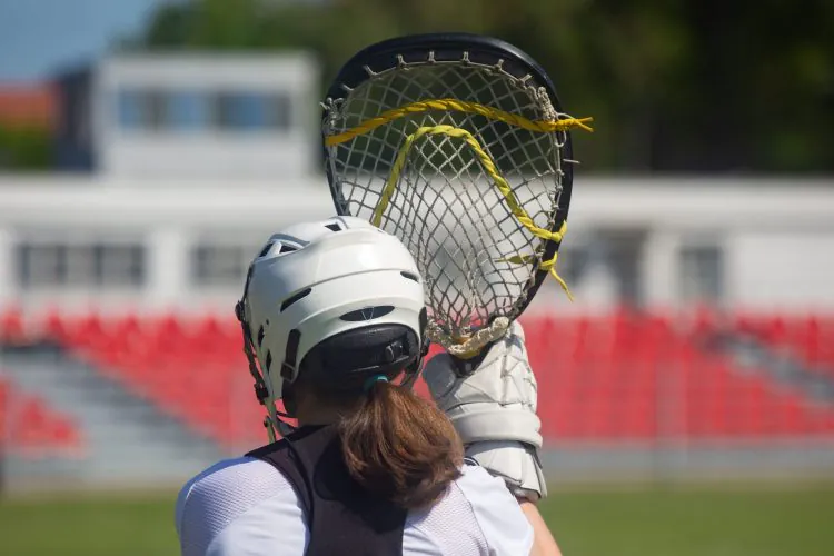 Rear view of a person in a white lacrosse helmet, holding a lacrosse stick with yellow lacing, on a field with stadium seating in the background.