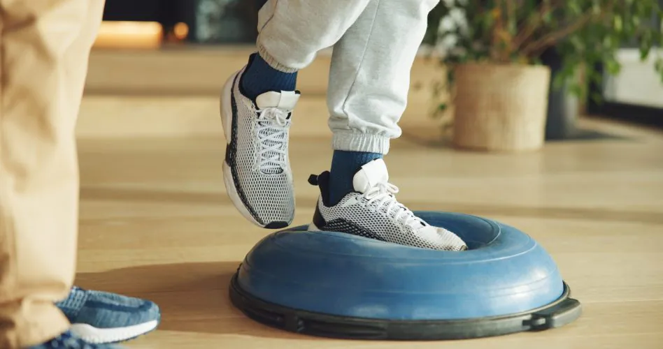 Feet, person and balance on ball for physiotherapy at house with assistance for mobility training. Physiotherapist, help and patient for stability exercise, muscle strength and rehabilitation session.