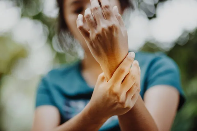 A person holds their wrist with their other hand, suggesting pain or discomfort, against a blurred green and white background.