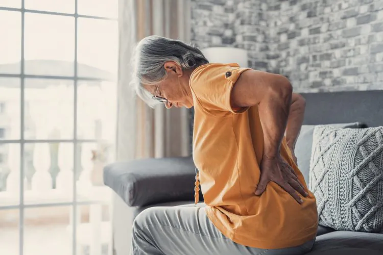 An elderly person sits on a couch, bending forward and holding their lower back with both hands, indicating back pain.
