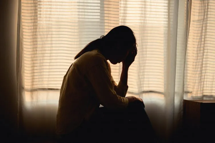 A silhouetted person sits with their head in their hand, facing a brightly lit window with sheer curtains.