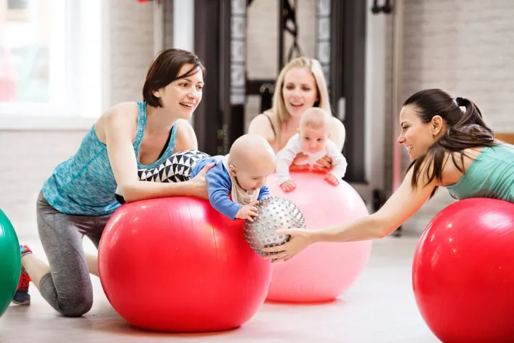 Three women and three babies interacting with colorful exercise balls and a silver spiky ball in a bright gym setting.